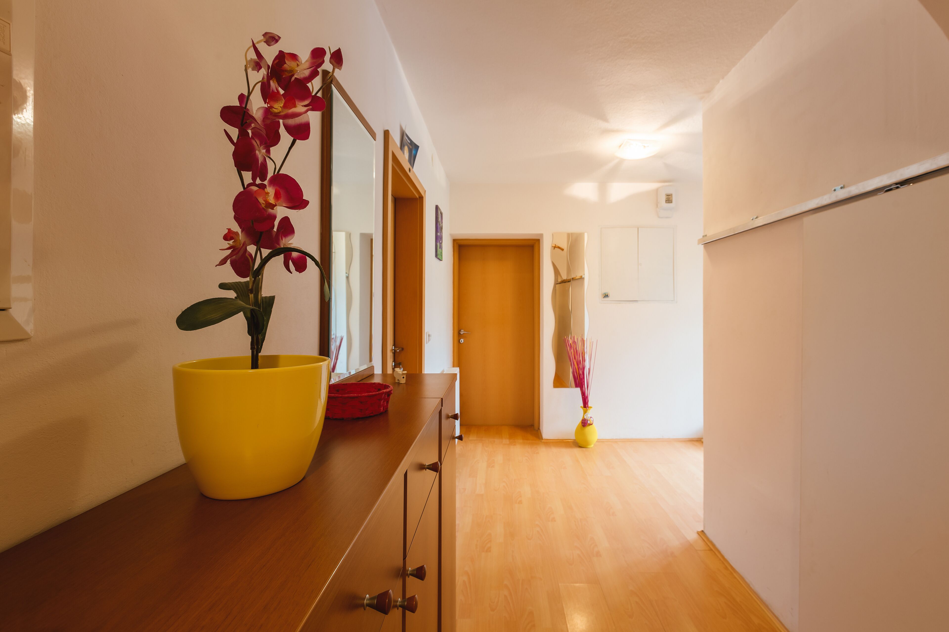 A hallway with wooden flooring, a yellow vase with red flowers, and a door at the end.