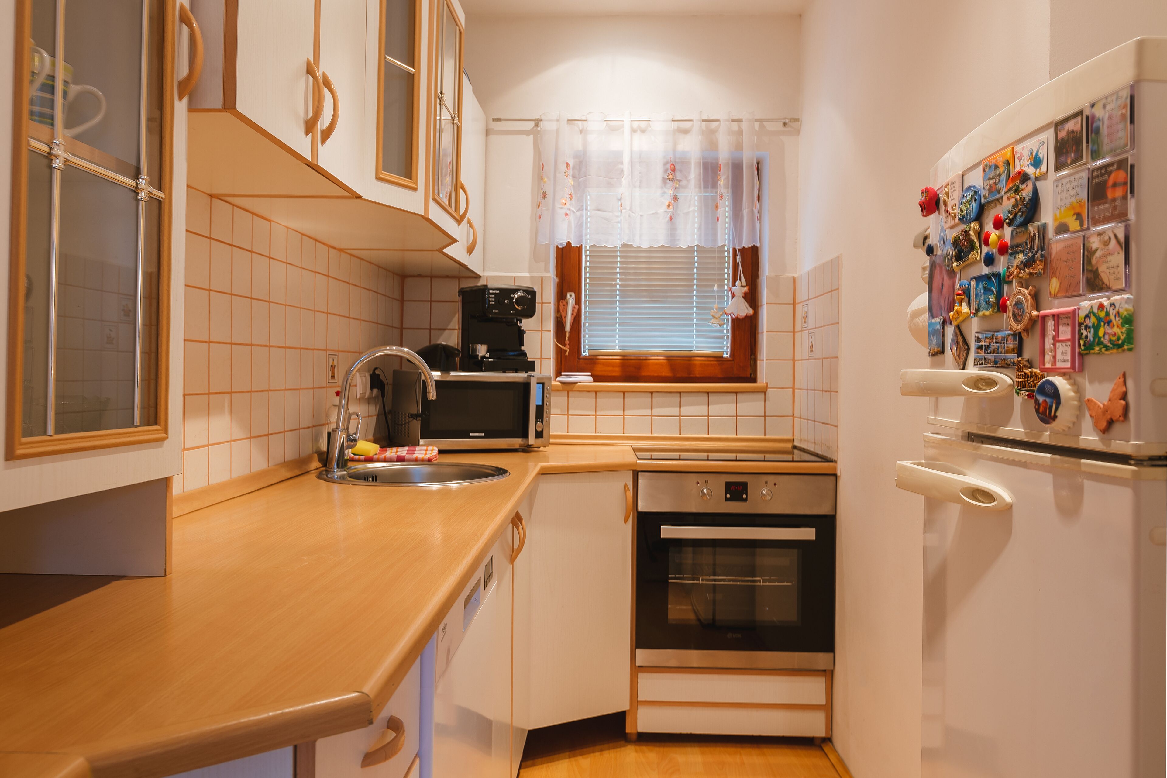 Cocina compacta con microondas, horno y lavavajillas. Se ve una ventana con persianas y cortina, y un refrigerador con imanes y fotos a la derecha.