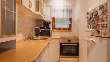 Cocina compacta con microondas, horno y lavavajillas. Se ve una ventana con persianas y cortina, y un refrigerador con imanes y fotos a la derecha.