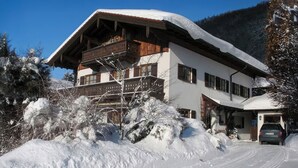 A two-story chalet with a wooden balcony, surrounded by snow-covered trees and a clear sky. - Apartment 54sqm, balcony, kitchen, 1 bedroom, living room, max. 2 persons (Ruhpolding)