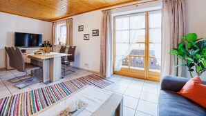 A modern living room with a wooden ceiling, a flat-screen TV, a dining area with a table and chairs, and a large window with curtains. - Apartment 54sqm, balcony, kitchen, 1 bedroom, living room, max. 2 persons (Ruhpolding)