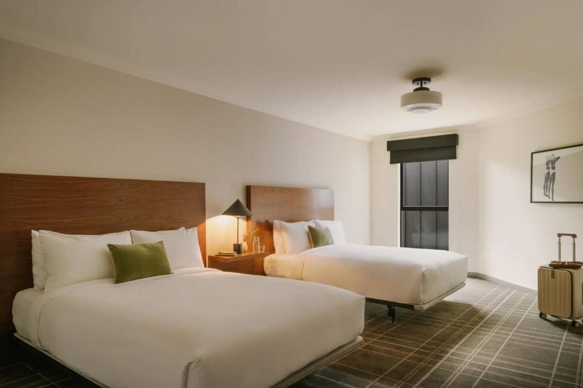 Habitación de hotel con dos camas, una mesita de noche con lámpara, una ventana con persianas y una maleta en el suelo.