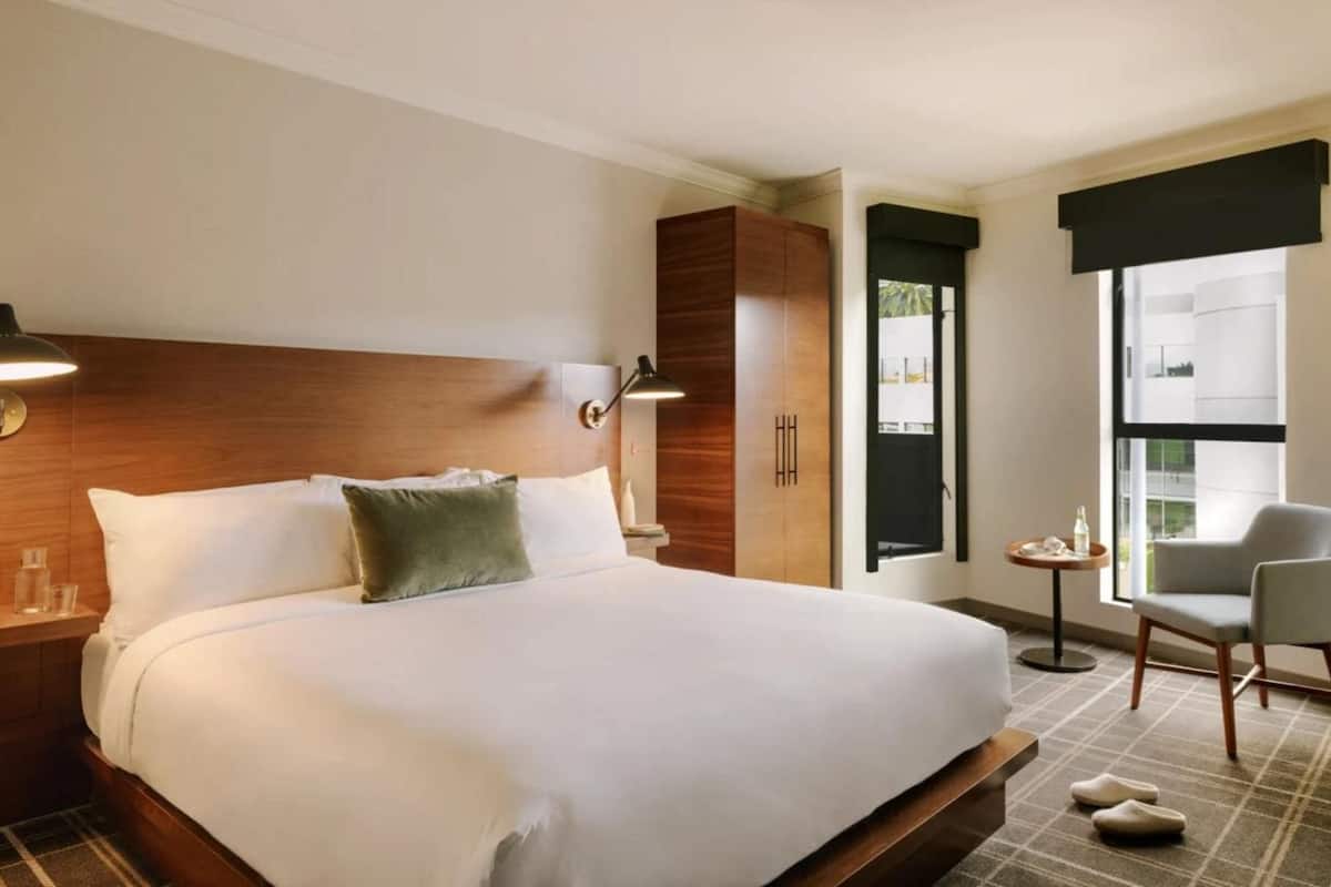 Habitación de hotel con una cama grande, una mesita de noche, una lámpara, una silla y una ventana con vistas.