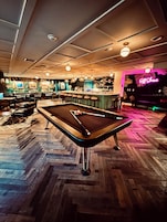 A pool table in a bar area with a neon sign reading "Off Shore" and a bar counter with stools.