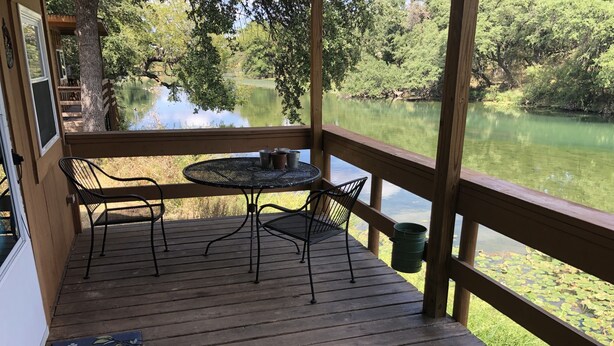 Una terraza de madera con una mesa y sillas da vista a un lago tranquilo, rodeado de árboles.
