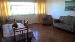 A living room with a patterned sofa, a patterned armchair, a dining table with a floral tablecloth, and a painting on the wall. - Maria Izabel (Salvador)