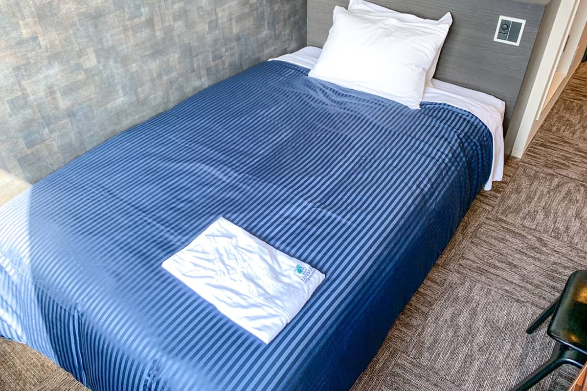 A neatly made bed with a blue bedspread and white pillows, a grey headboard, and a small bedside table with a clock and a sign.