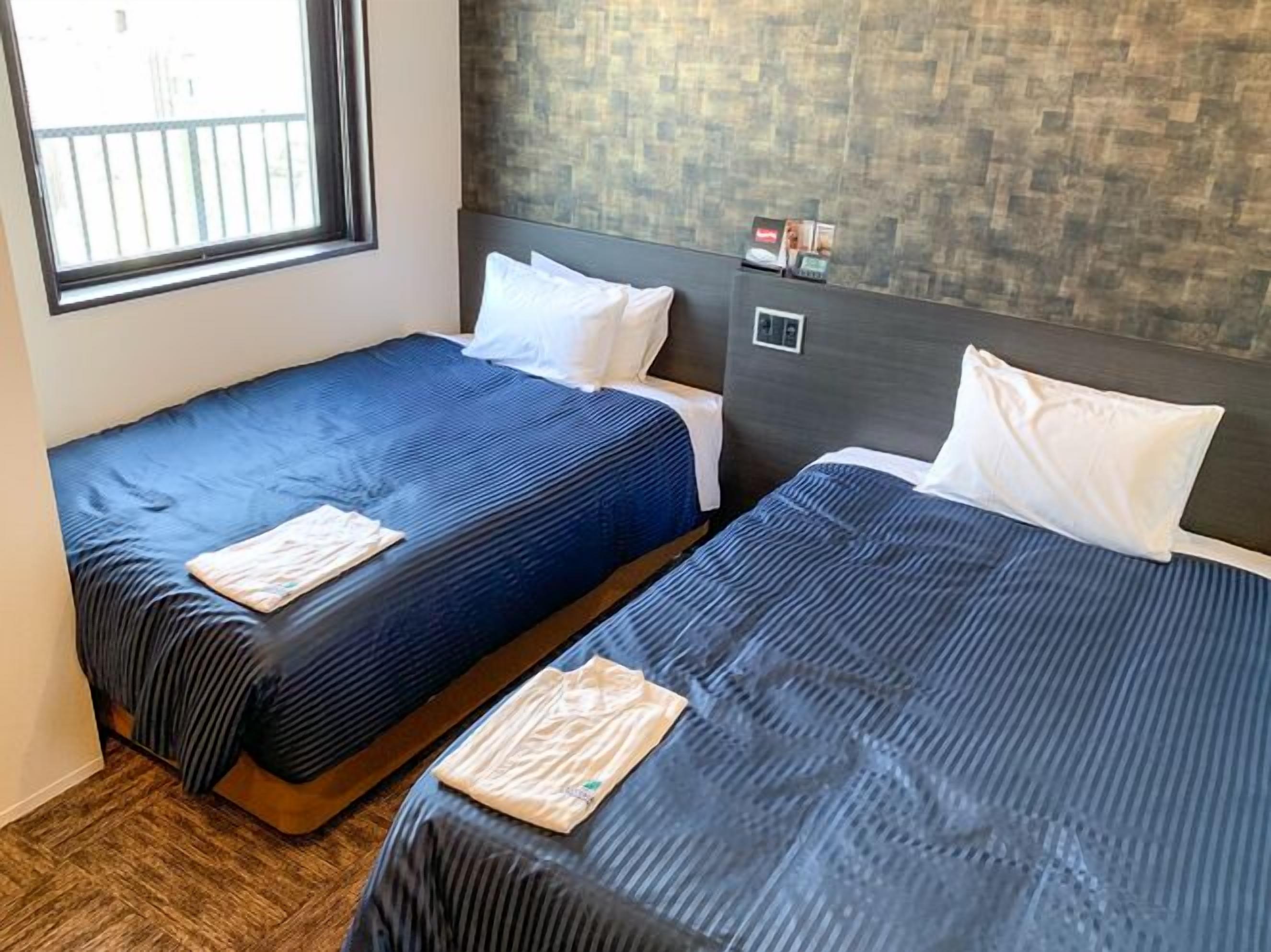 A hotel room with two beds, a window, a brick-patterned wall, and a small control panel.