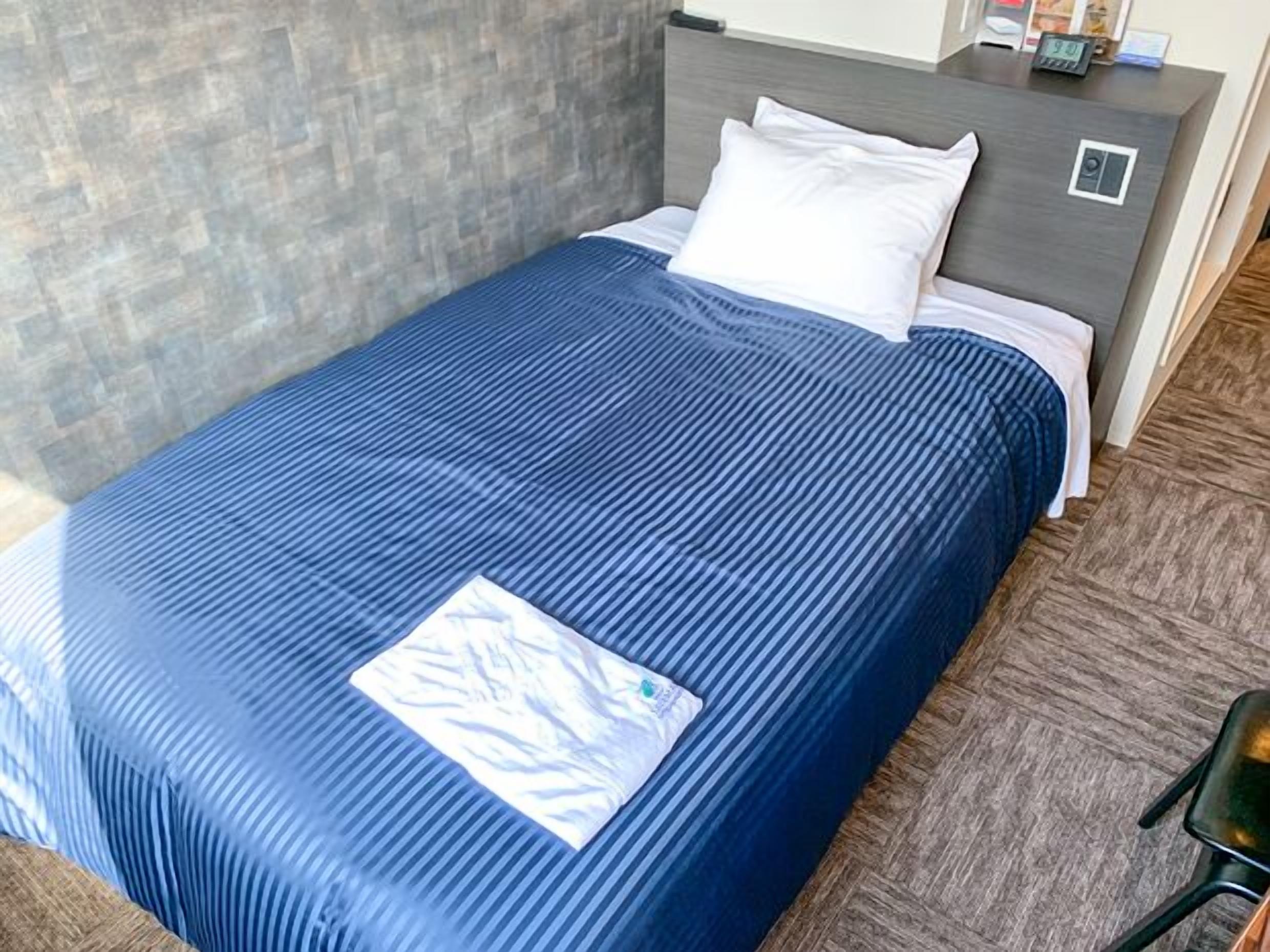 A neatly made bed with a blue bedspread and white pillows, a grey headboard, and a small bedside table with a clock and a phone.