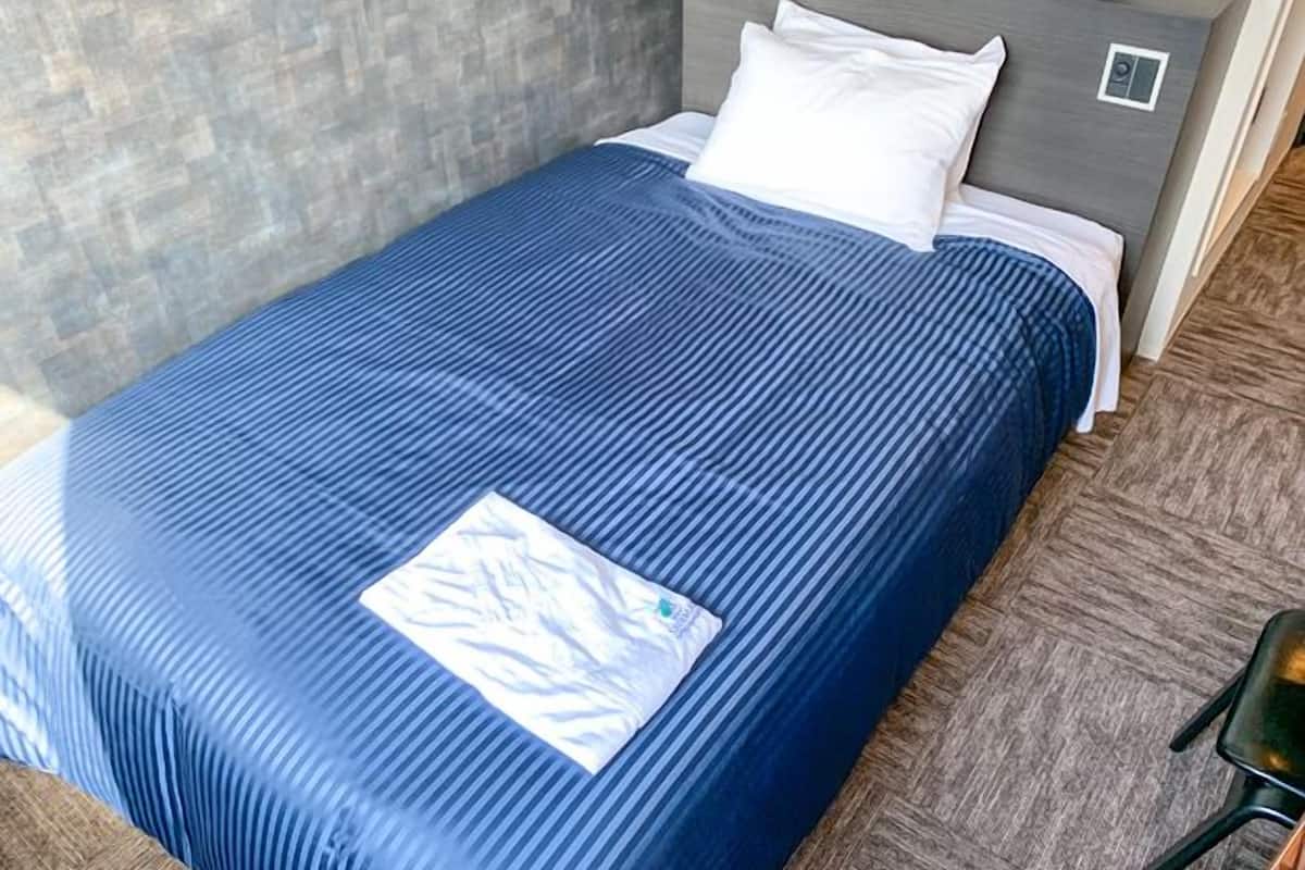 A neatly made bed with a blue bedspread and white pillows, a grey headboard, and a small bedside table with a clock and a phone.