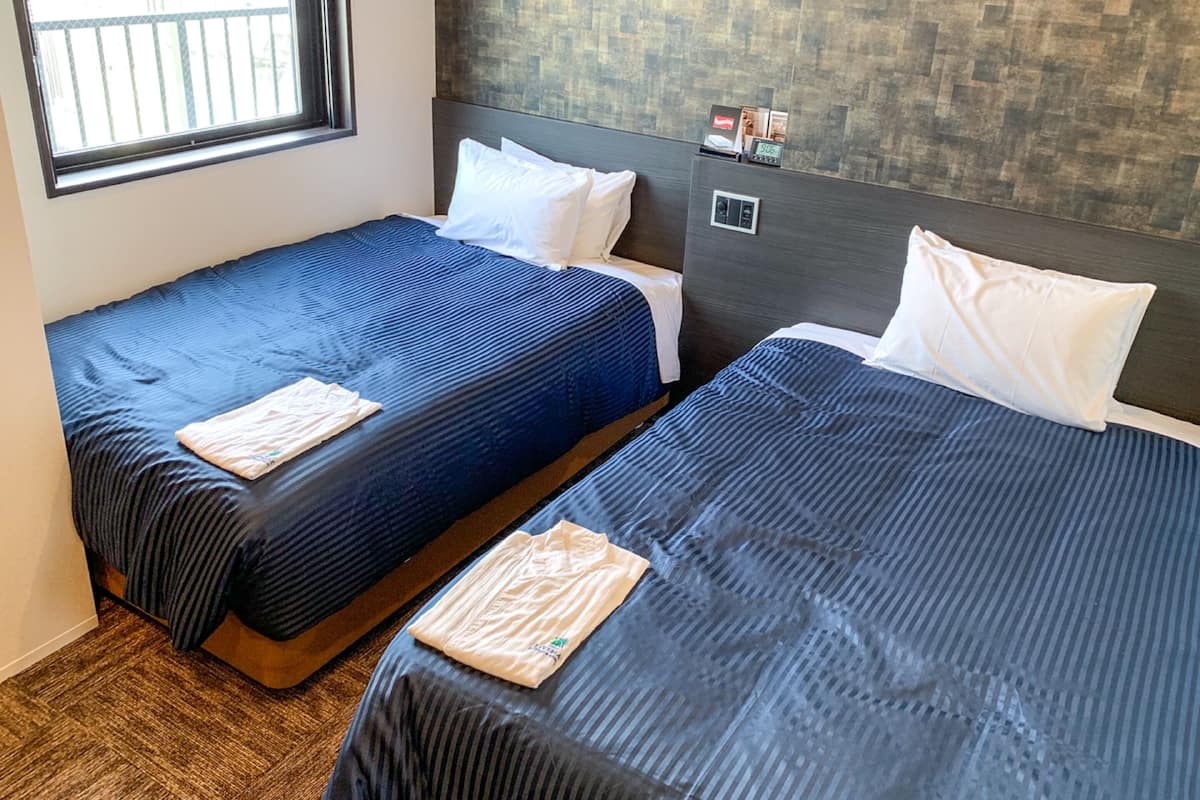A hotel room with two beds, each with a blue bedspread and white pillows. There is a window with a view of a balcony, and a wall-mounted control panel.