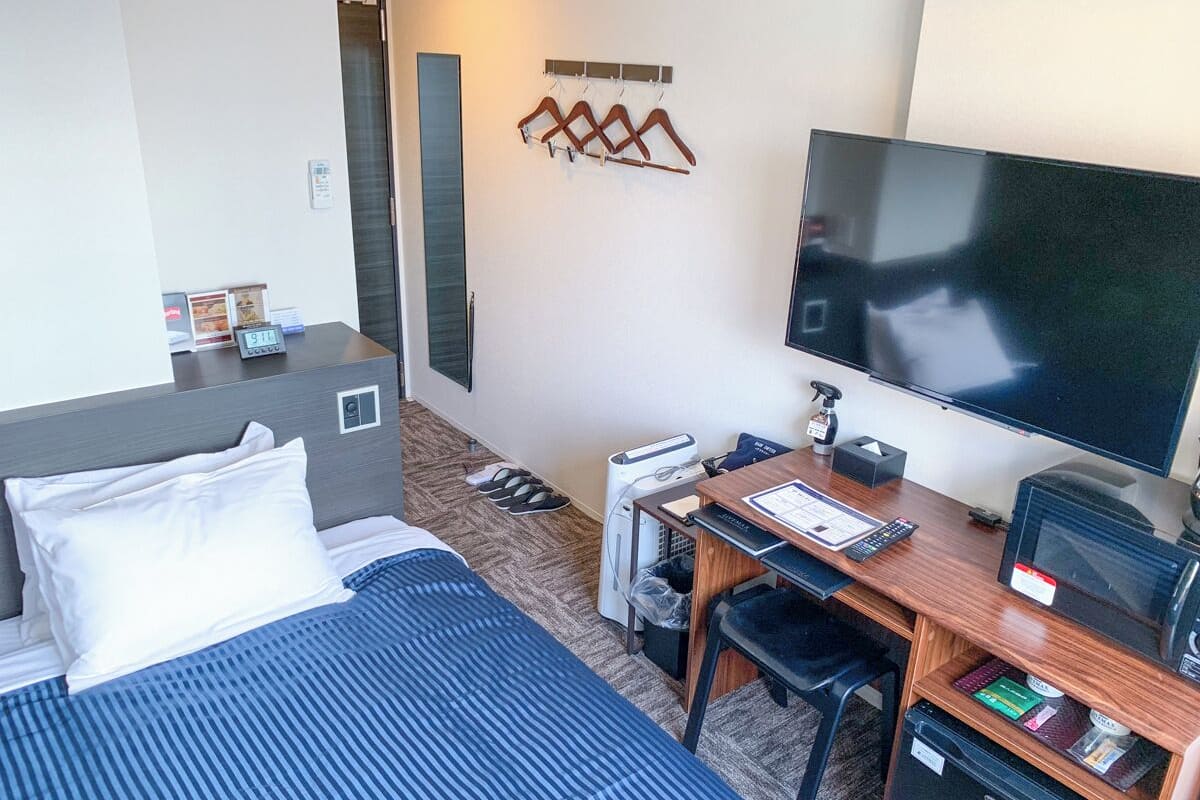 A hotel room with a bed, desk, television, and a wall-mounted coat rack.