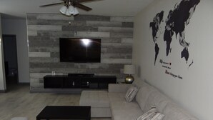 Ein modernes Wohnzimmer mit einem großen Flachbildfernseher an einer Holzwand, einem grauen Sofa, einem schwarzen Couchtisch und einer Weltkarte als Wandbild.