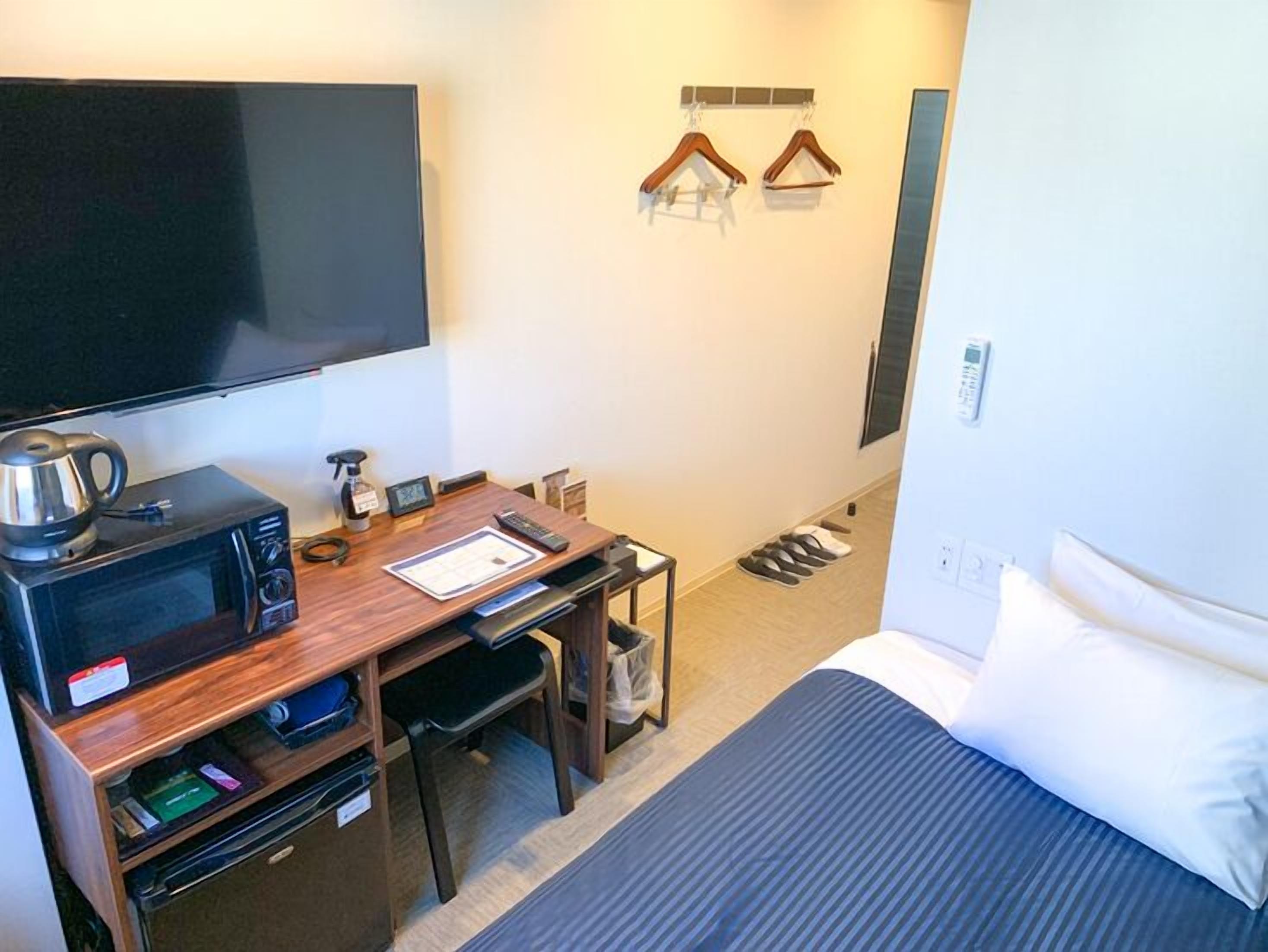 A hotel room with a bed, a desk with a microwave, a TV, and a shoe rack.