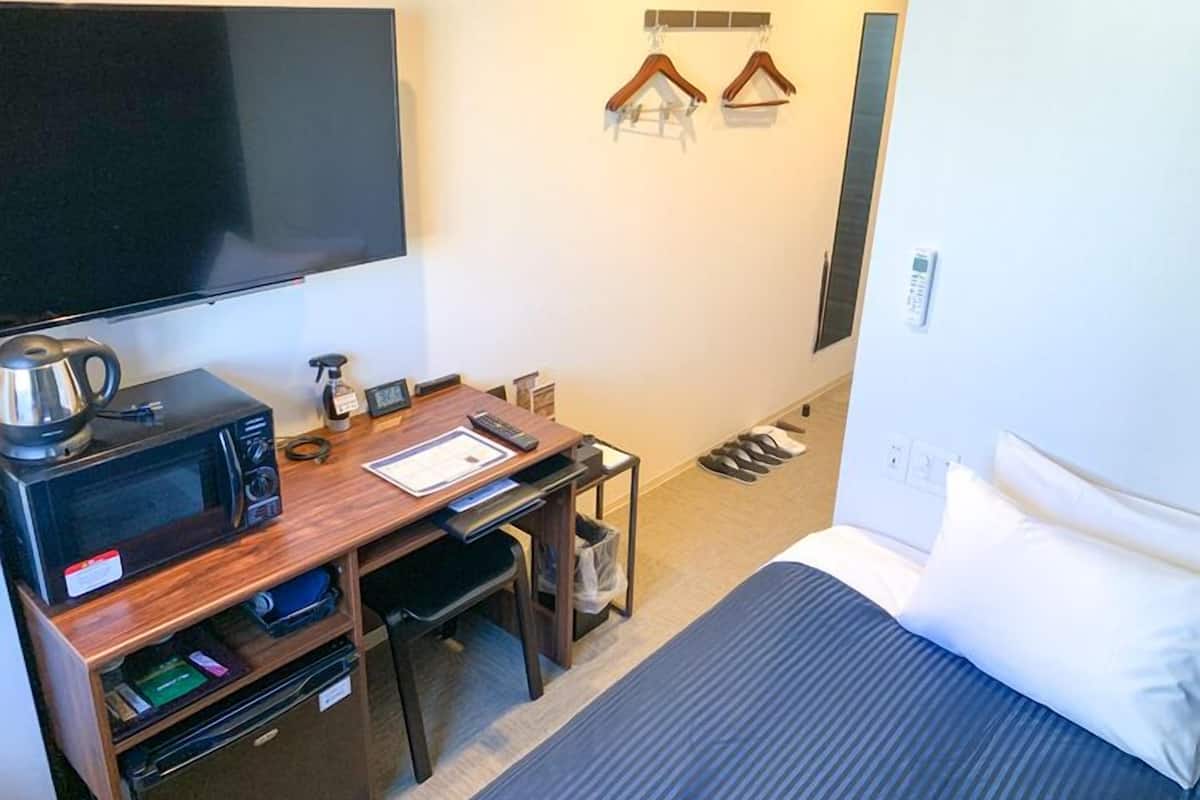 A hotel room with a bed, a desk with a microwave, a TV, and a shoe rack.