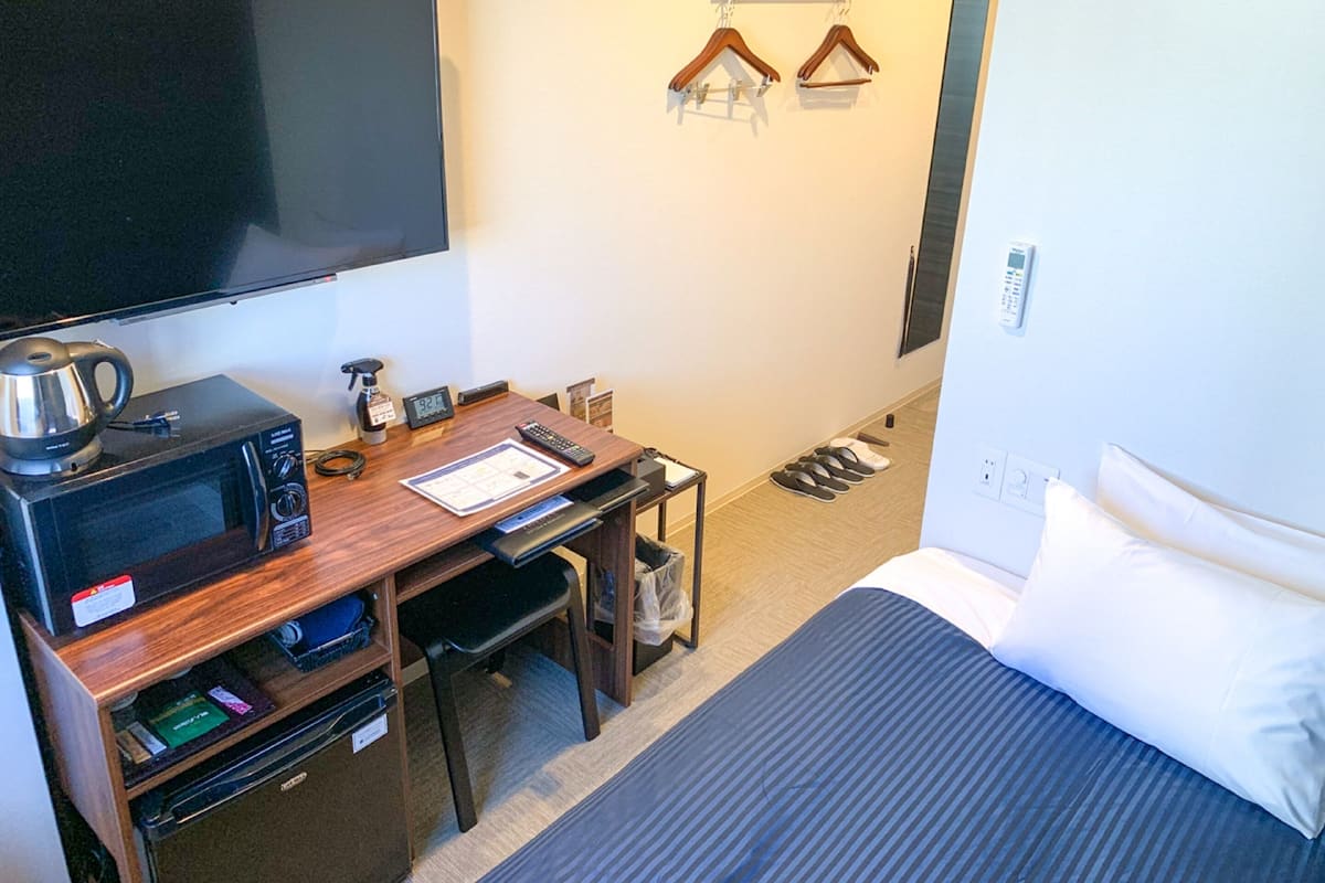 A hotel room with a bed, a desk with a microwave, a kettle, a clock, and a TV.