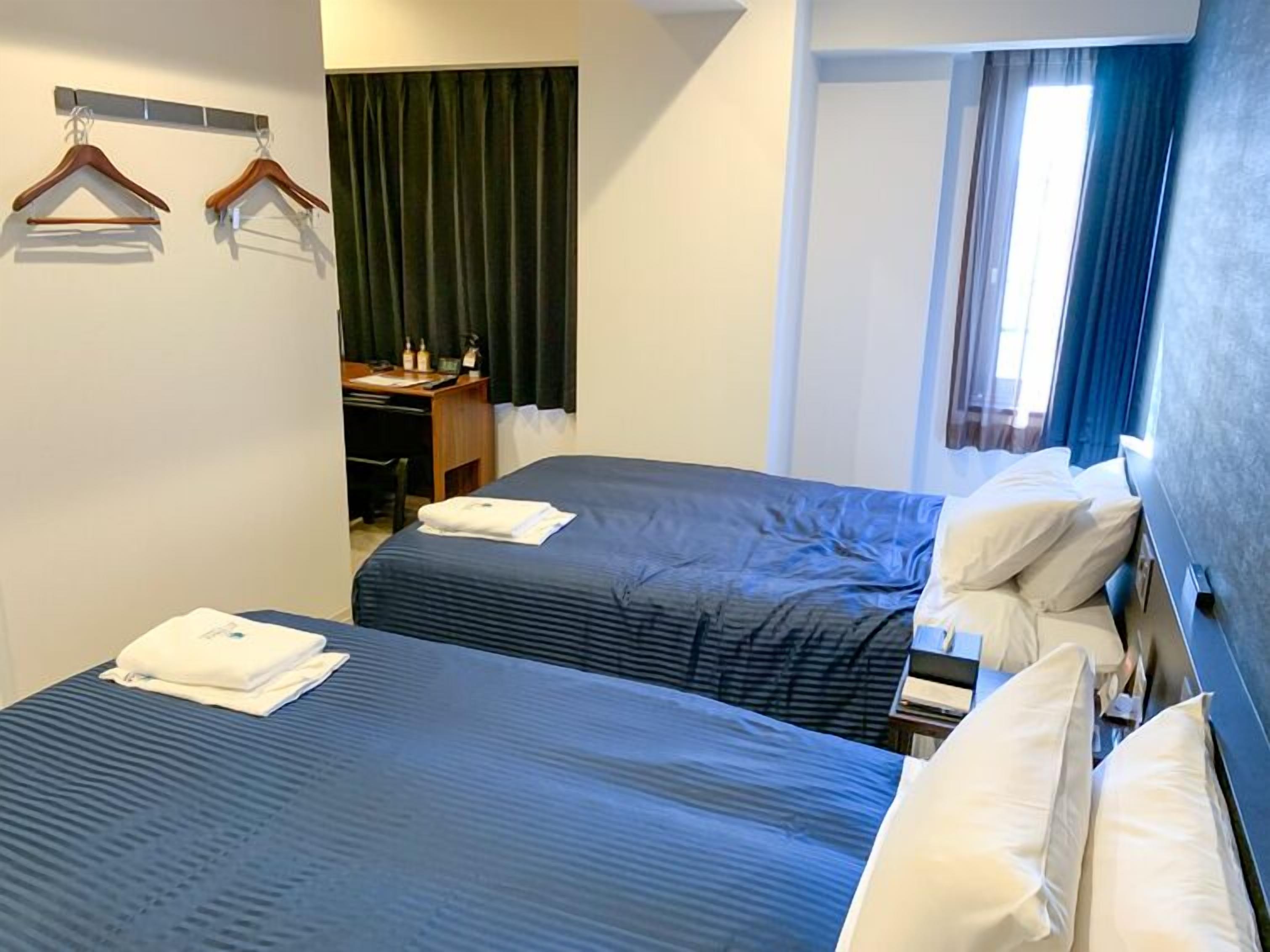 A hotel room with two beds, a desk with a chair, a coat rack, and a window with curtains.