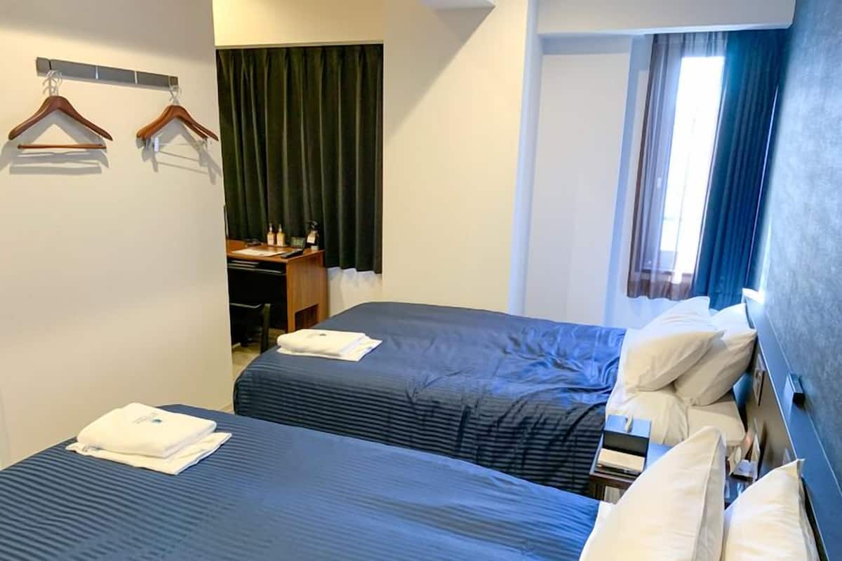 A hotel room with two beds, a desk with a chair, a coat rack, and a window with curtains.
