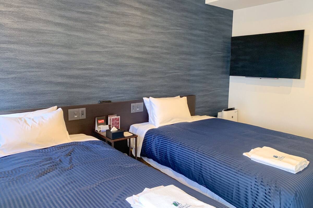 A hotel room with two beds, a flat-screen TV mounted on the wall, and a small table with a lamp.