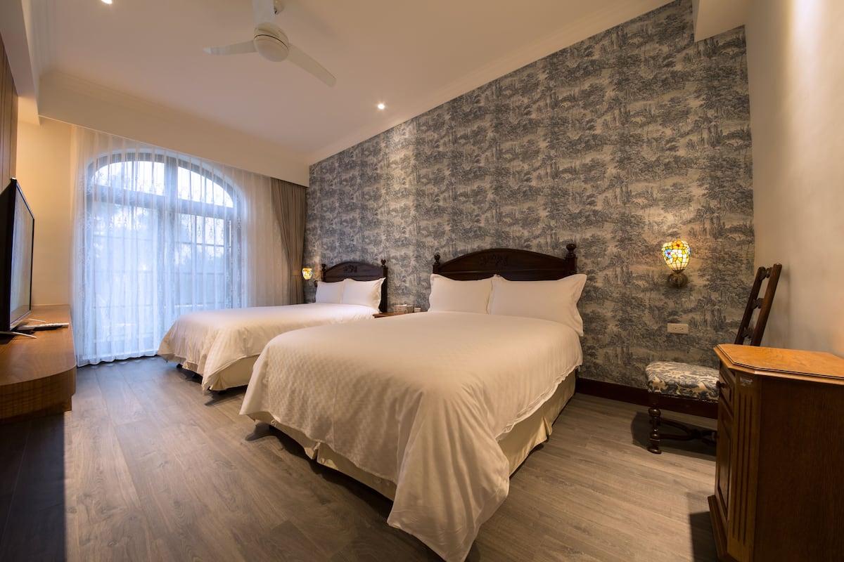 A hotel room with two beds, a television, a desk, and a large window with curtains.