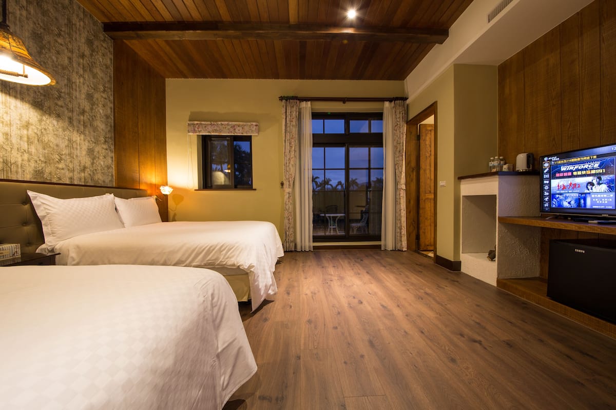 A hotel room with two beds, a television, a wooden ceiling, and a view through a glass door.