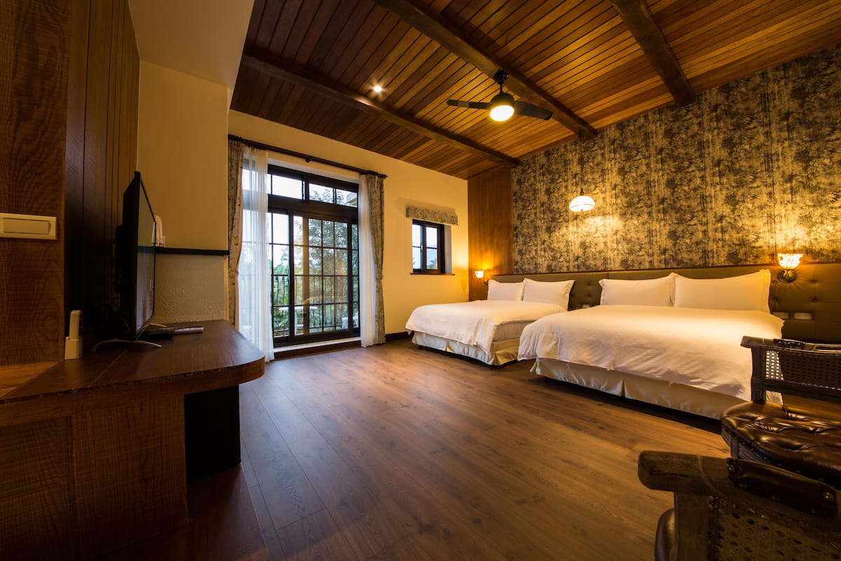 A hotel room with a wooden ceiling, two beds, a desk, and a large window with curtains.
