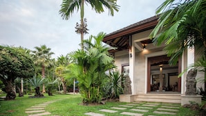 A tropical garden with a stone pathway leading to a building with a tiled roof and a stone statue.