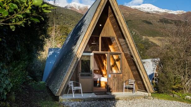Una baita di legno a forma triangolare con porta in vetro, un letto e una sedia all'interno, immersa in un paesaggio montuoso.