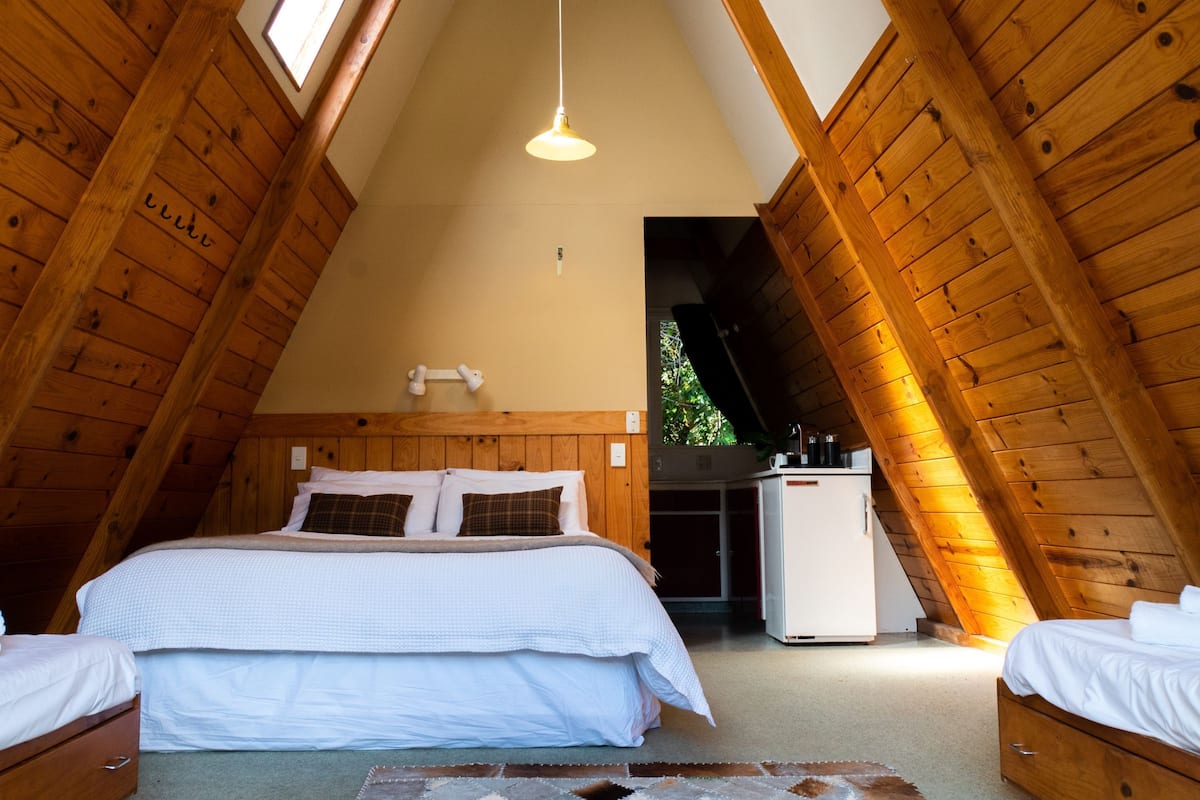 A cozy attic bedroom with a wooden ceiling, a bed with white bedding, a small white refrigerator, and a hanging light fixture.