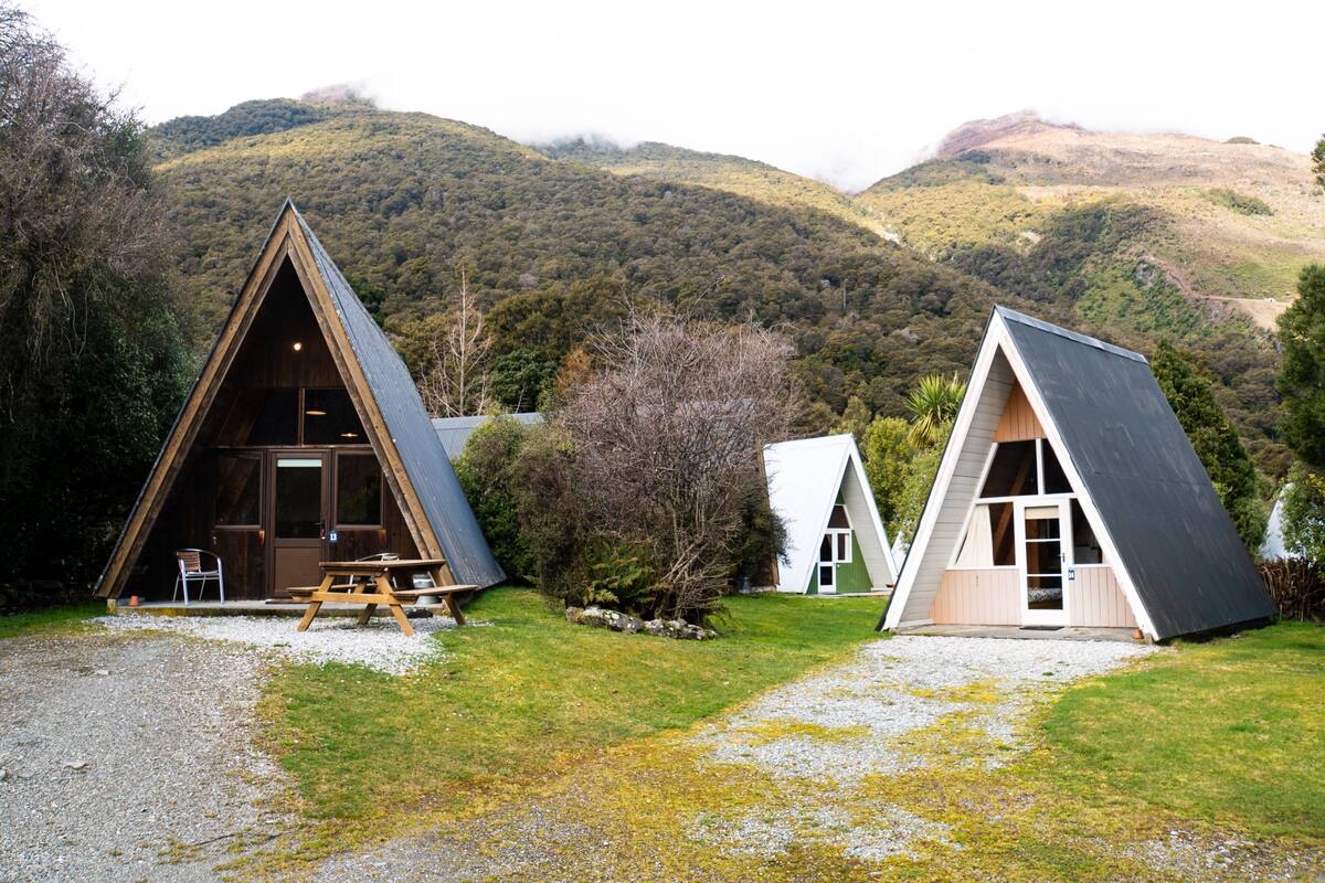 Two A-frame cabins with wooden exteriors, set in a grassy area with a mountainous backdrop.