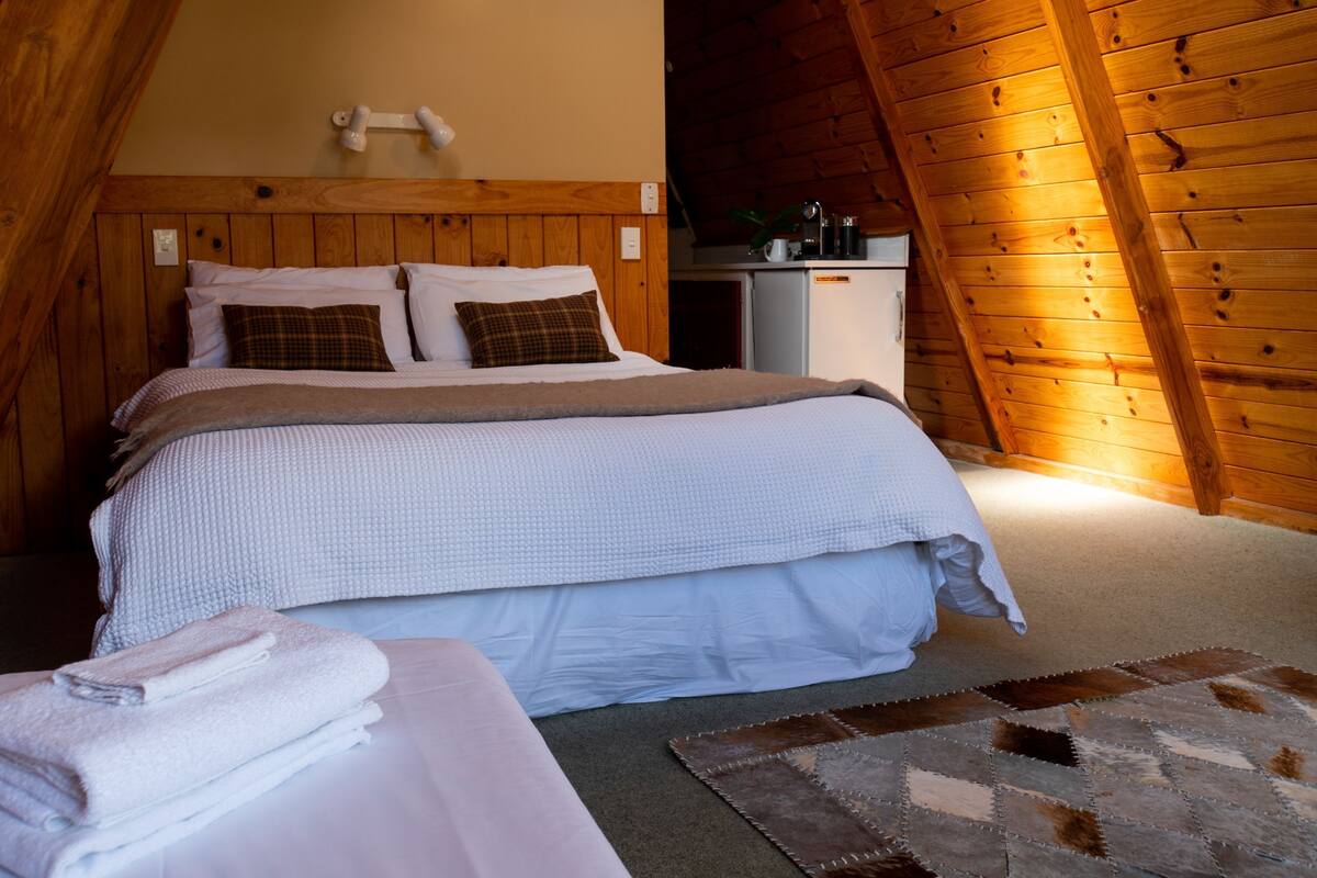 A cozy bedroom with a wooden ceiling, a bed with white bedding, a small bedside table, and a nightstand.