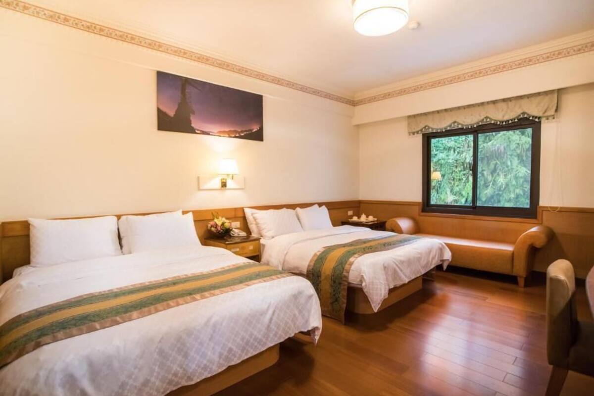 A hotel room with two beds, a nightstand with a lamp, a bench, a window with a view of greenery, and a painting on the wall.