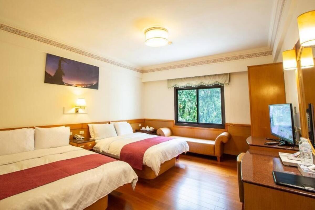 A hotel room with two beds, a desk, a chair, a TV, and a window with a view of greenery.