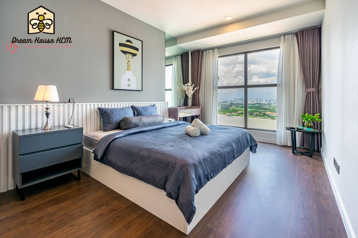 Un dormitorio moderno con una cama grande, una mesita de noche, una lámpara y vistas a la ciudad a través de amplias ventanas.