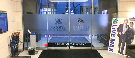 A hotel entrance with a glass door, a black mat, a bench, and a sign with 'LIVE MAX' and 'LIVE GROUP' logos.