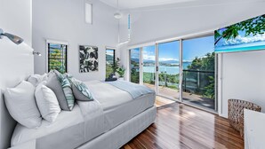 A modern bedroom with a large bed, a view of the sea through floor-to-ceiling windows, and a balcony with a view.