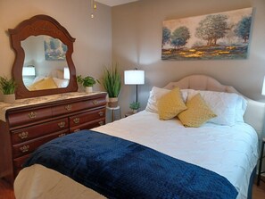 A bedroom with a large bed, a dresser with drawers, a mirror, a painting on the wall, and a lamp.