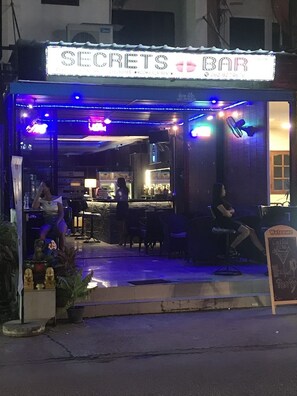 Un bar appelé « SECRETS BAR », avec une enseigne au néon, des tables à l’extérieur et un menu écrit à la craie.