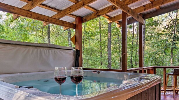Un jacuzzi con dos copas de vino tinto, rodeado por una terraza de madera y vistas al bosque.