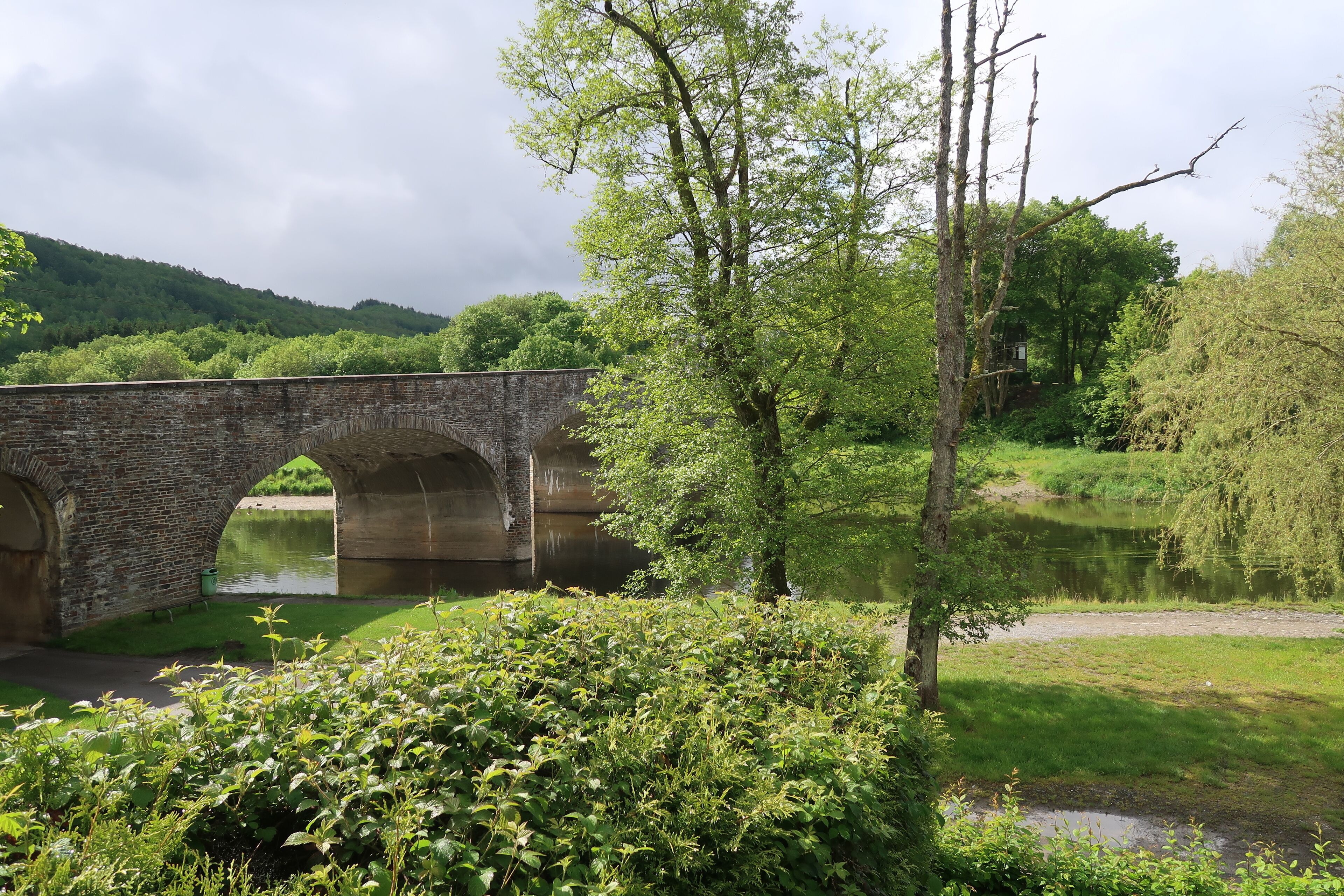 A stone bridge over a calm river, surrounded by lush greenery and trees.