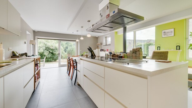 Une cuisine moderne dotée d’un grand îlot central, de meubles blancs et offrant une vue sur la verdure environnante.