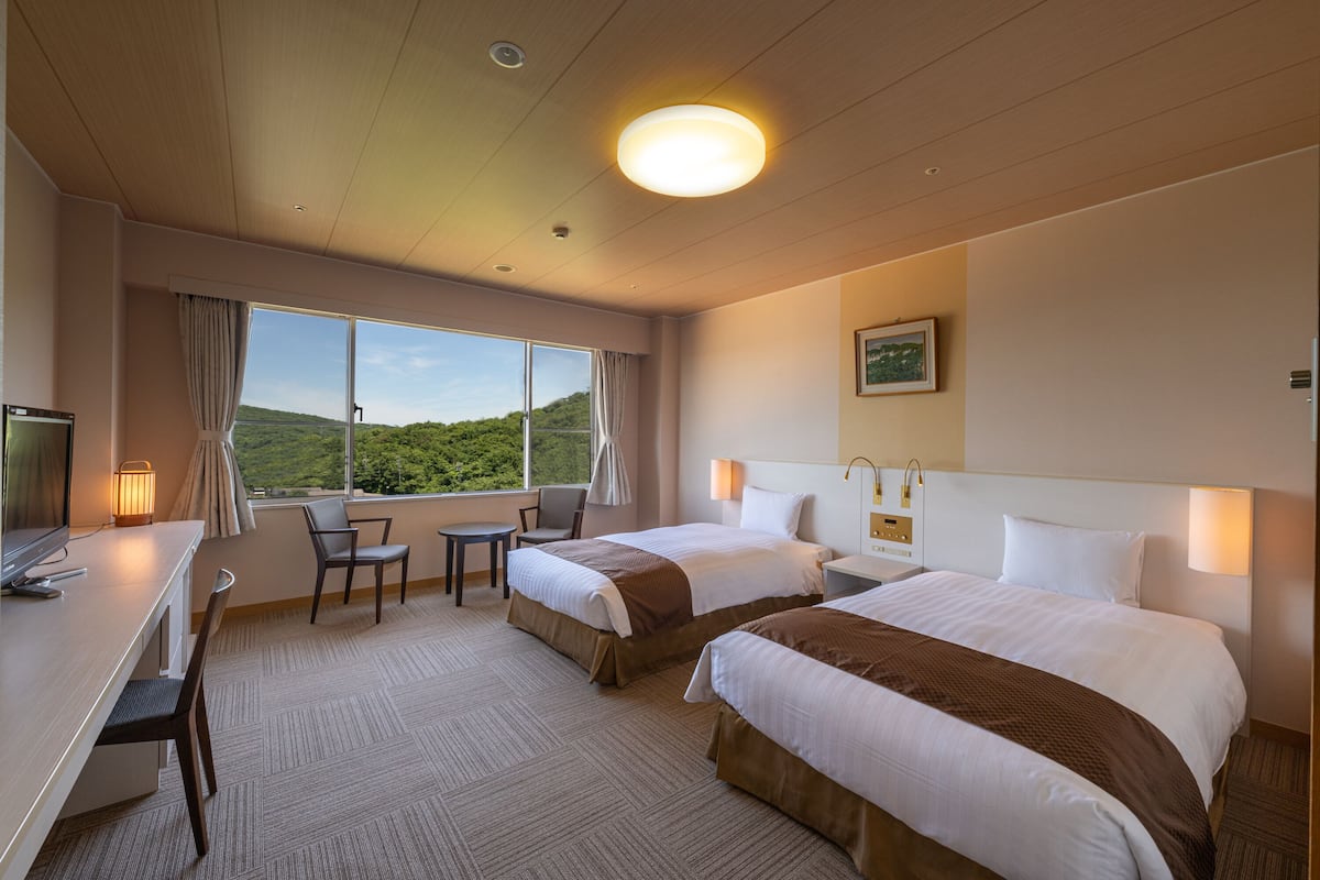 Habitación de hotel con dos camas, un escritorio, una silla, una mesita y una ventana con vistas a zonas verdes.