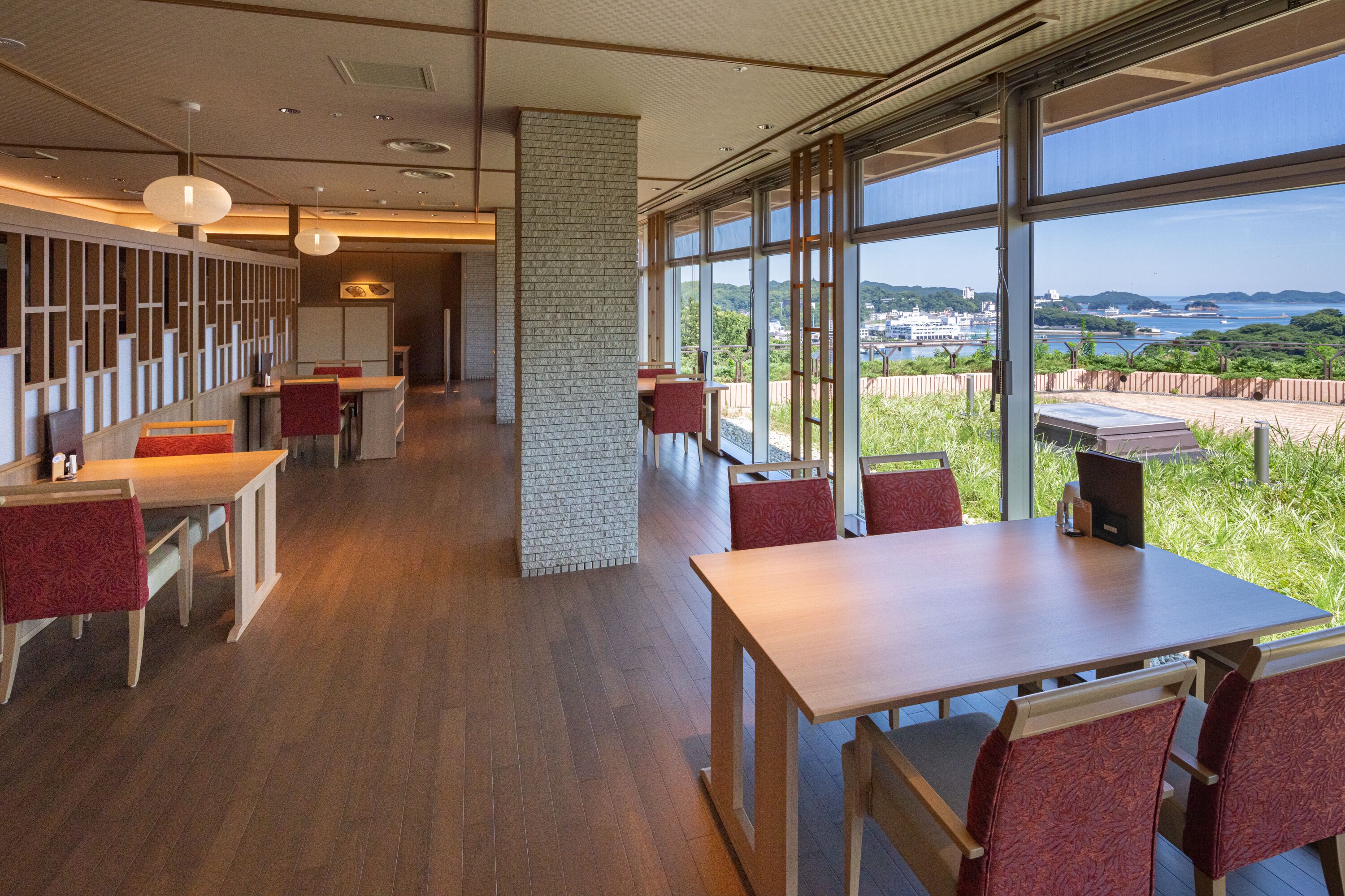 A modern dining area with wooden tables and red upholstered chairs, large windows offering a view of the sea and distant hills.