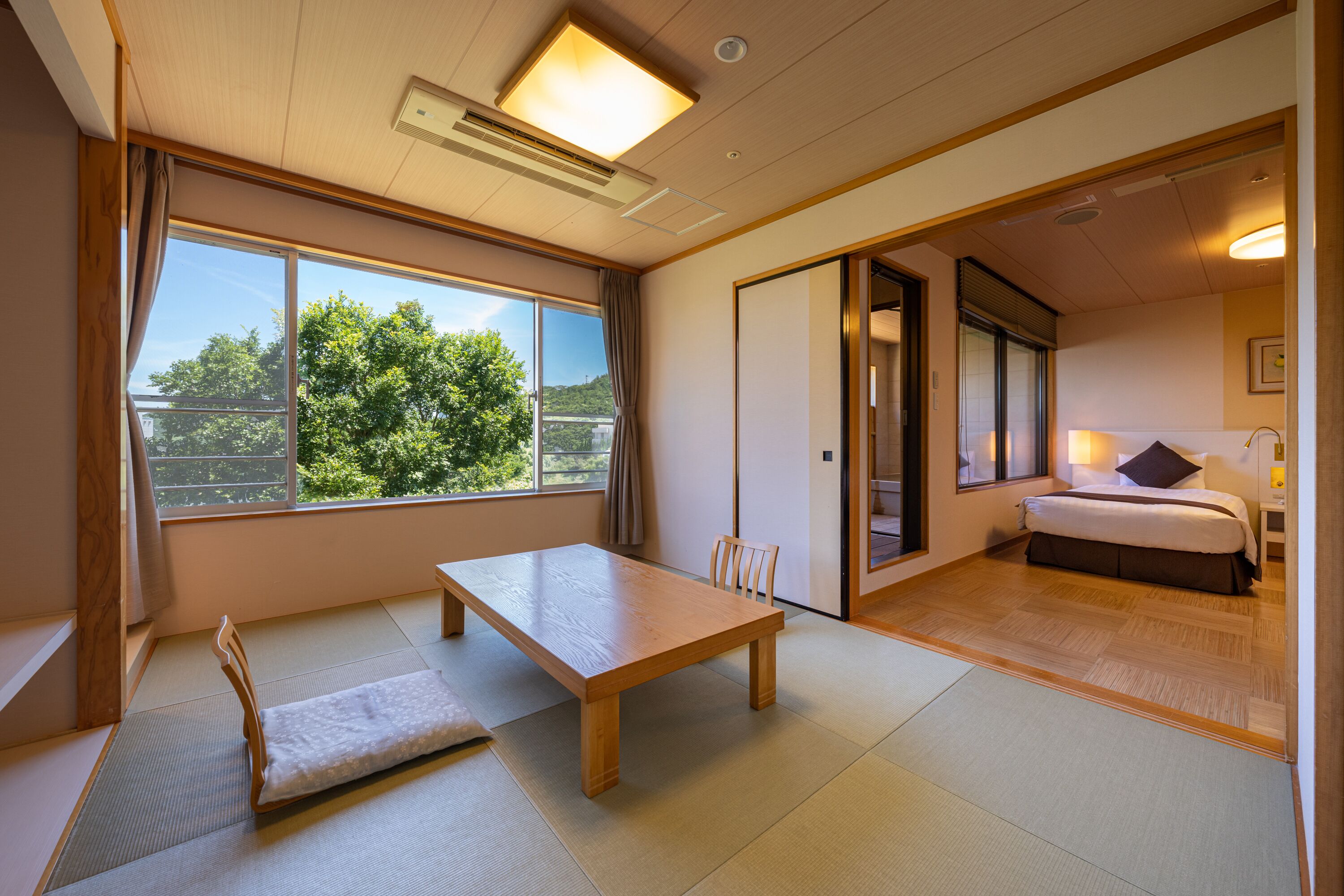 A room with tatami flooring, a wooden table, a bed, and a view of greenery outside.