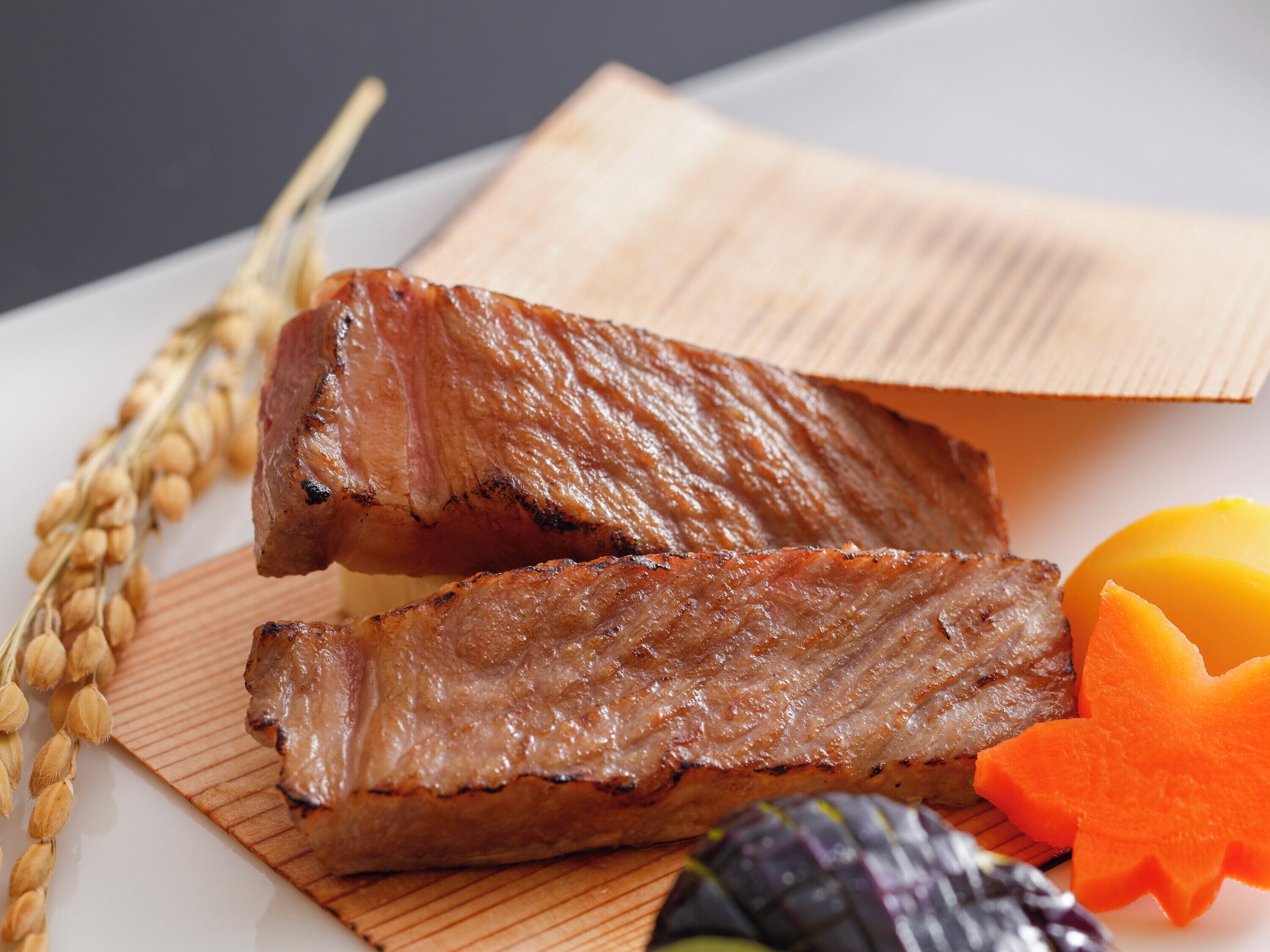 A wooden cutting board with grilled meat, a bunch of wheat, and a carved pumpkin.