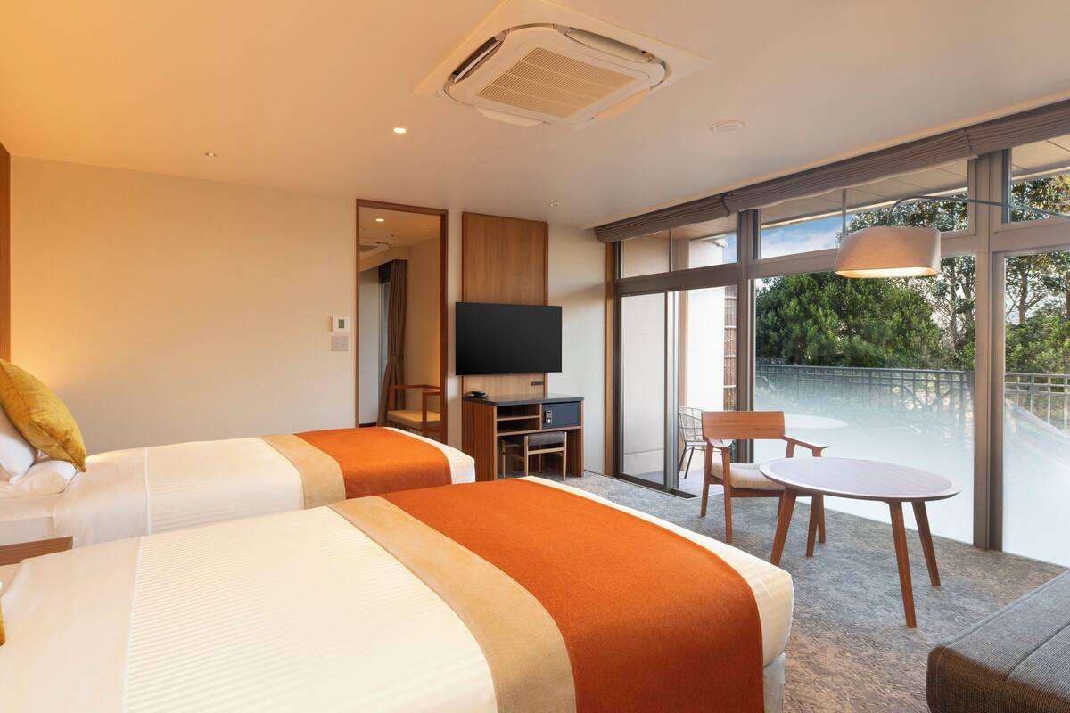 Habitación de hotel con dos camas, televisión, escritorio y balcón con mesa y sillas.