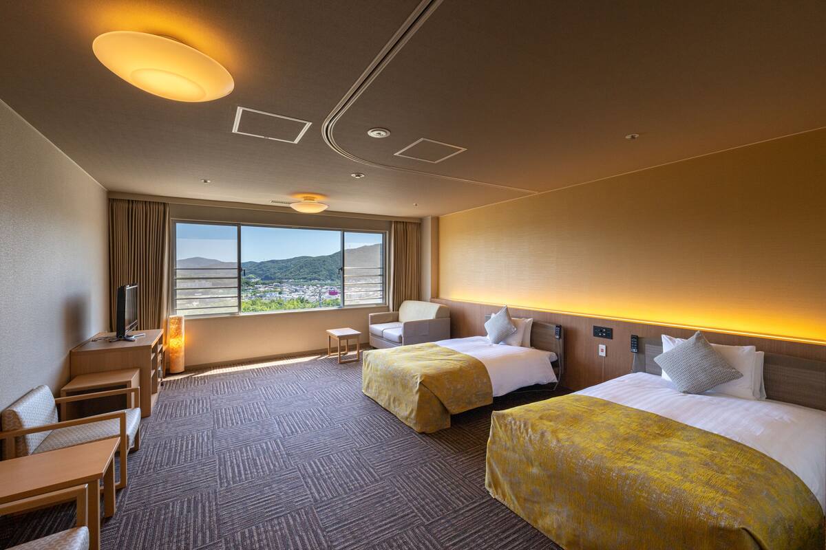 Habitación de hotel con dos camas, un escritorio, una silla, un televisor y un ventanal con vistas a montañas.