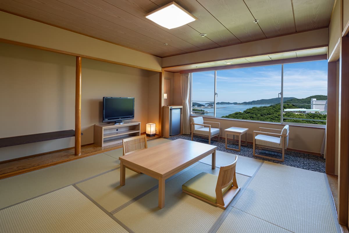 Una habitación de hotel moderna con piso de madera, asientos de estera de tatami, televisor de pantalla plana, un refrigerador pequeño y un ventanal grande que ofrece una vista panorámica del mar y las colinas.