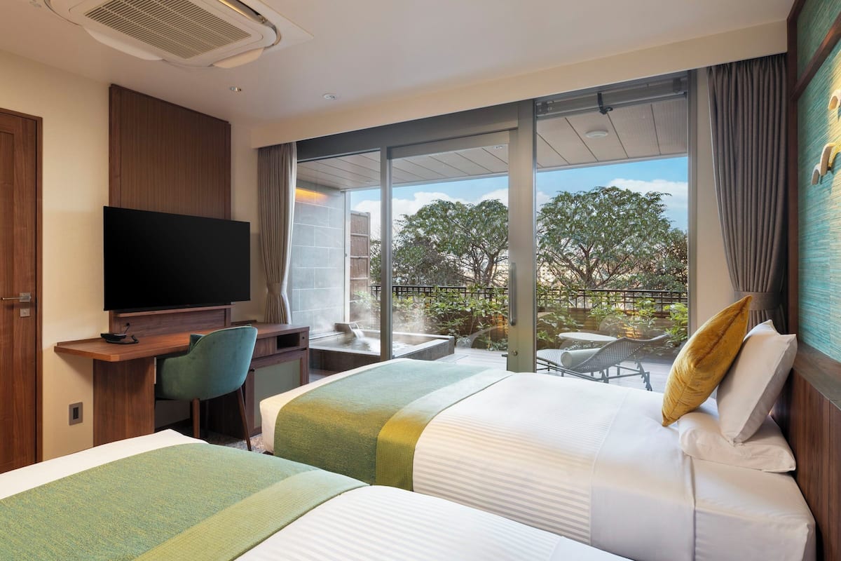 Habitación de hotel con dos camas, un escritorio con televisor y un balcón con vistas a árboles y una piscina.