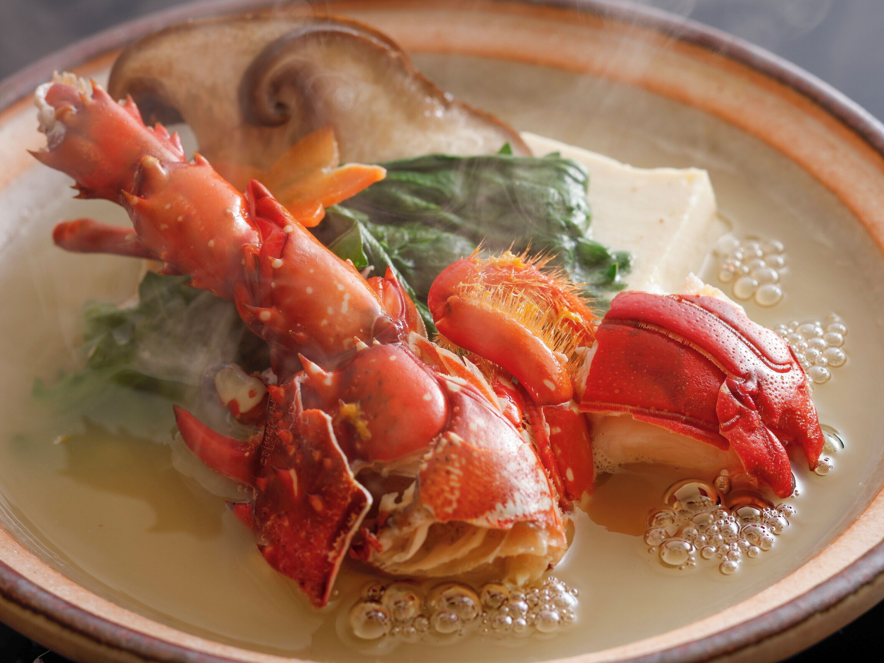 A bowl of seafood soup with a lobster, mushrooms, and greens.